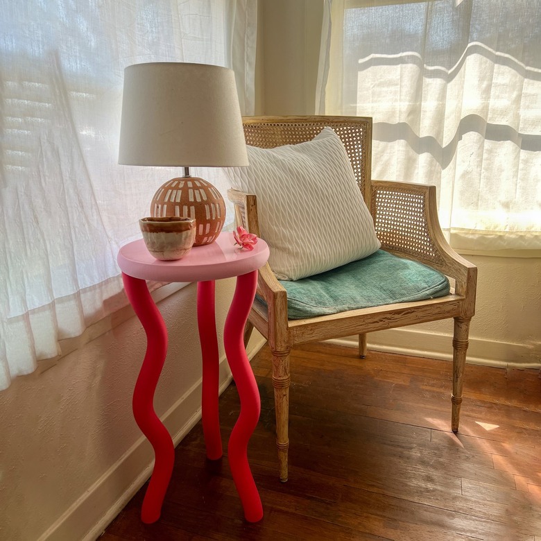 The image shows a corner of a brightly lit room. A side table is arranged next to a wicker chair. The side table has three squiggly hot pink legs and a circular bubblegum pink surface. A desert-colored ceramic mug and a round terracotta lamp are placed on the side table.
