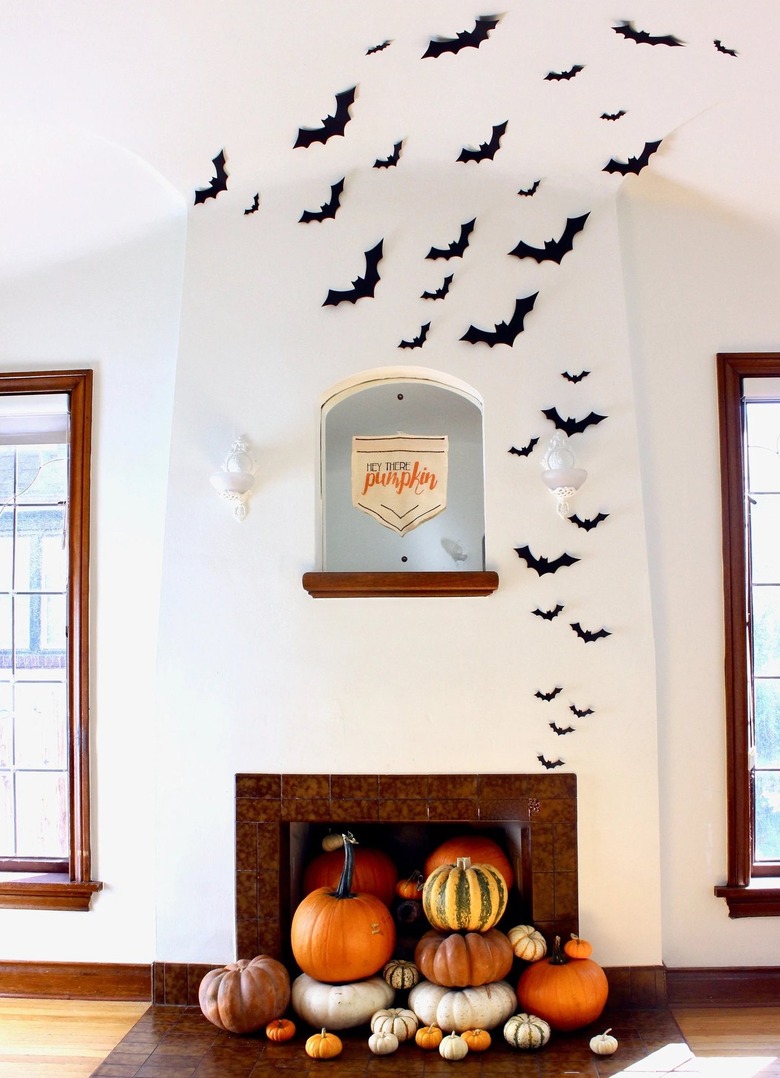 A white fireplace with paper bats that look like they are flying out of it. The hearth is filled with pumpkins of different shapes