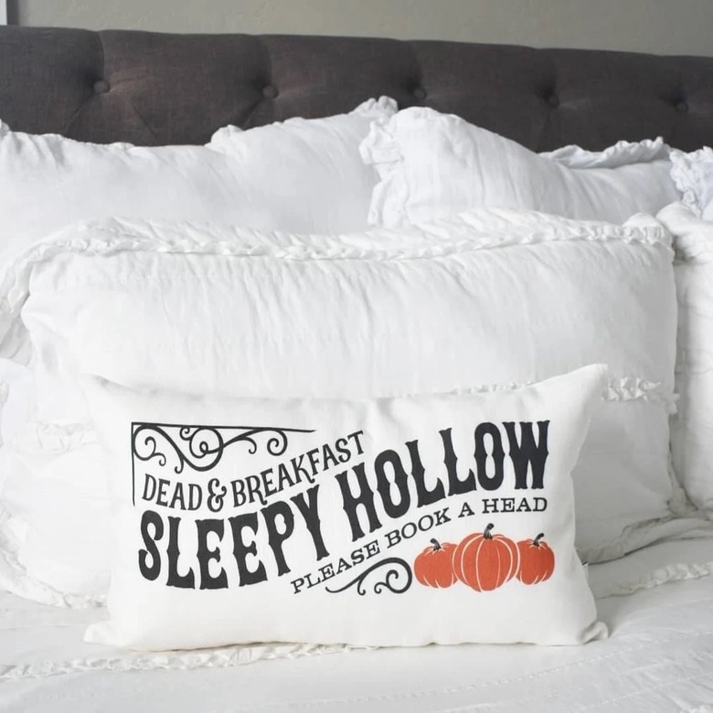 A bed covered in white pillows. The front throw pillow says Dead and Breakfast. Sleepy Hollow. Please book a head. Below