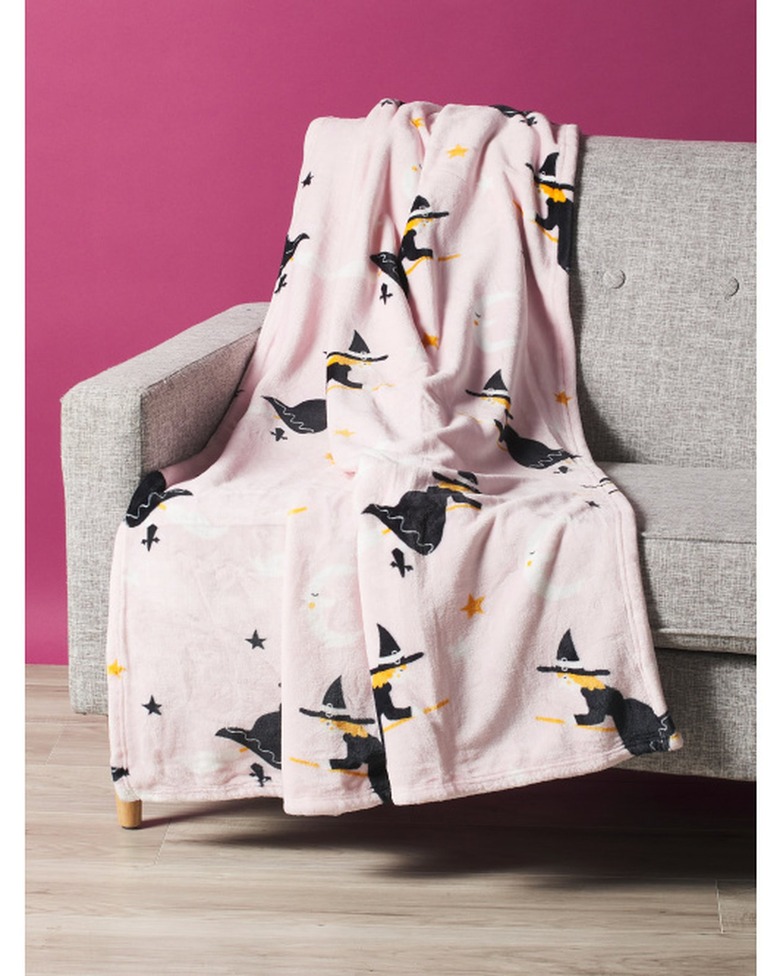 A pink fluffy throw blanket on a light grey fabric couch in front of a magenta wall. The pink blanket has flying witches