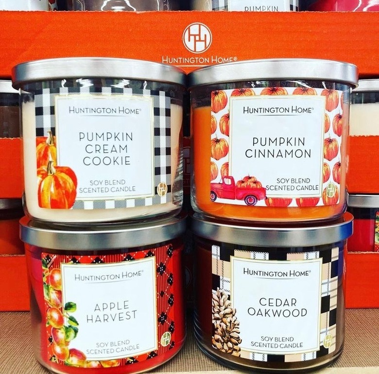 A close up of four seasonal Aldi candles. The Pumpkin Cream Cookie label has a black gingham background and pumpkins in the corner. The Pumpkin Cinnamon label has a pumpkin printed label with a small red truck in the corner. Apple Harvest has a red plaid label with apples on a branch on the left side