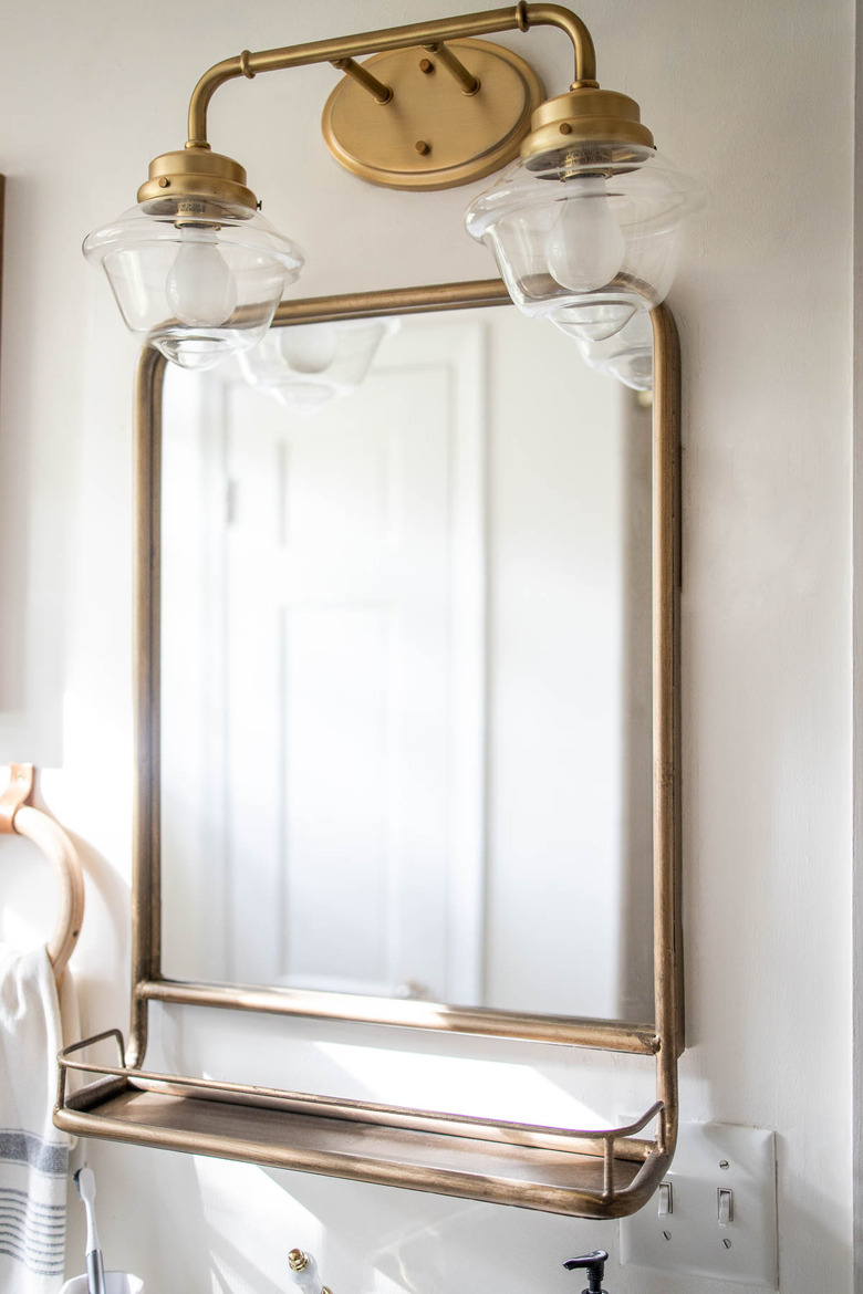 Brass two-arm lighting fixture above brass pharmacy-style mirror in bathroom