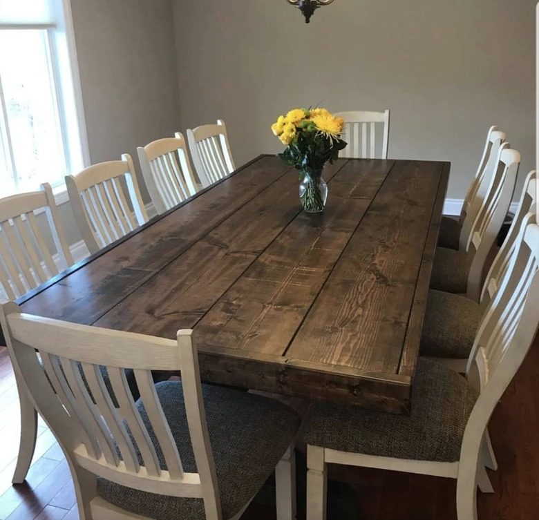 Image of a large dark farmhouse table surrounded by ten white rake-back chairs. There's a vase with yellow flowers on the table.