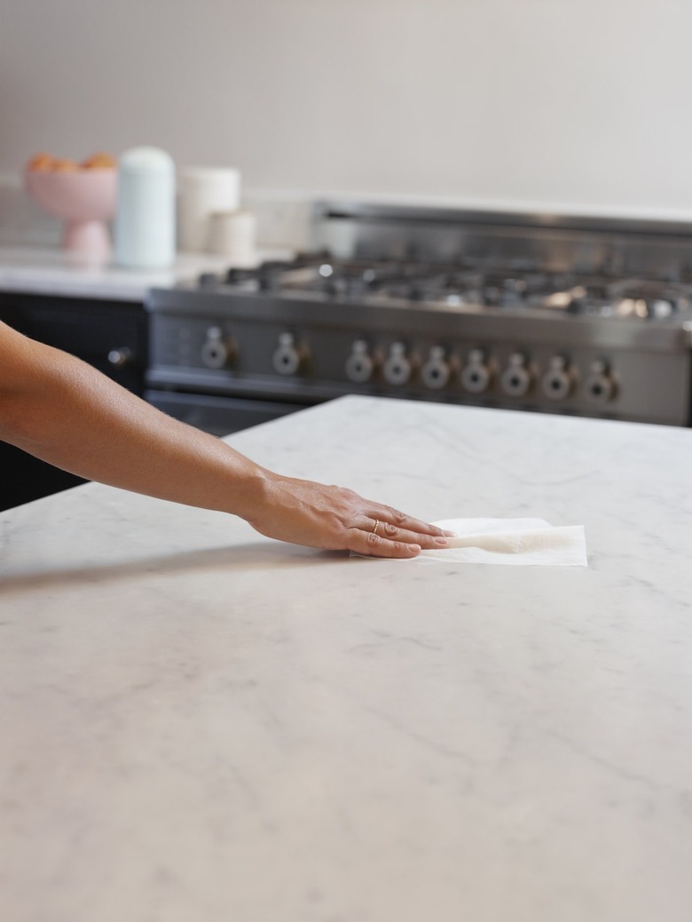 A medium-skin toned hand wipes down a marble kitchen counter with a wipe. In the background there's a stove top