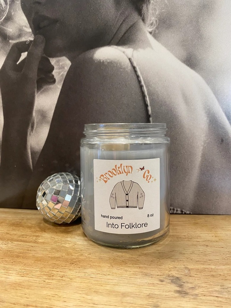 A small Brooklyn + Co candle rests on a wooden surface next to aa mini disco ball and in front of a Taylor Swift Folklore vinyl. The candle features a label with Taylor Swift's white cardigan on the front