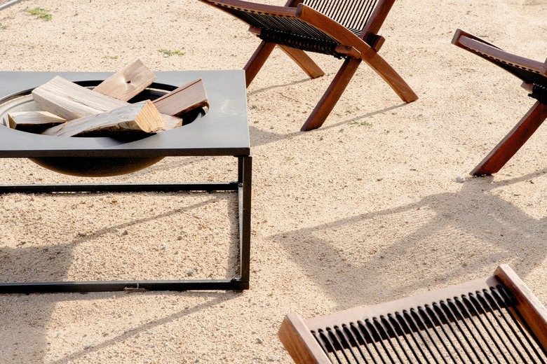 Close up of a black metal fire pit with cut logs in a dirt patio area. Parts of wooden patio chairs are visible.