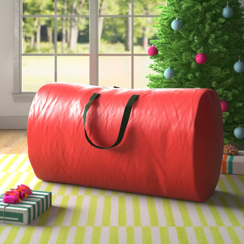 Red Christmas tree storage bag with black handles on rug next to Christmas tree