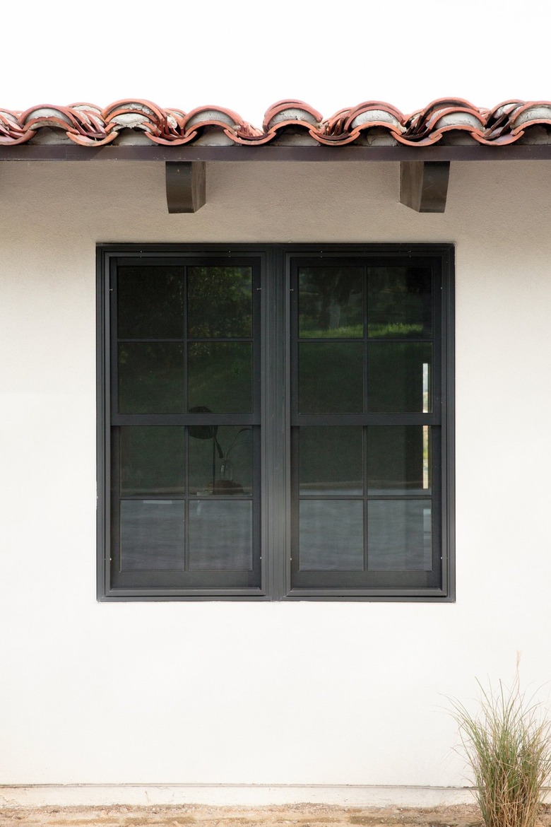 Side of a Spanish-style home with white walls and a clay tile roof with dark wood support beams. There is a rectangular window in the middle of the wall. On the ground