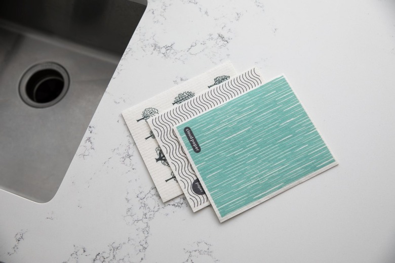 Three swish cloths rest in a pile on a white marble kitchen counter next to a sink. The cloth on top is teal with thin white lines throughout