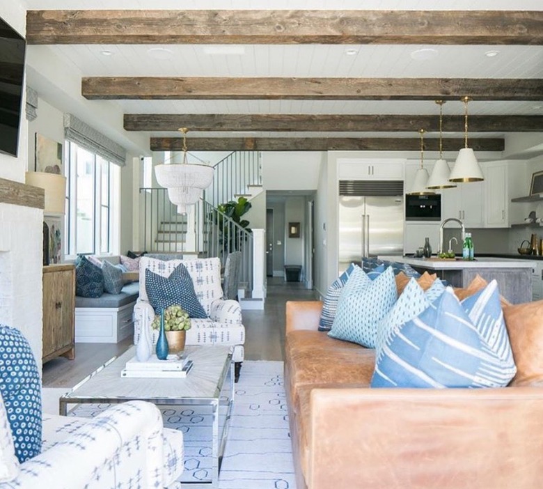 Open family room/kitchen with caramel leather couch with blue and white throw pillows and side chairs and wood-beamed ceilings.