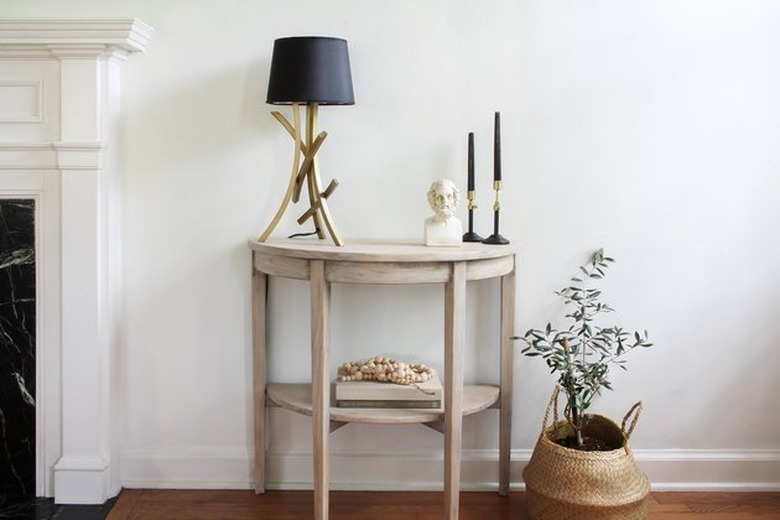 Bleached wood console table with black lamp and candles on top next to a basket with plant inside.