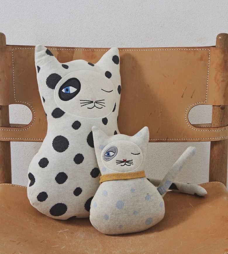 A white cat pillow with black cats. One eye is closed and the other is open with a black dot around it. Next to the large pillow