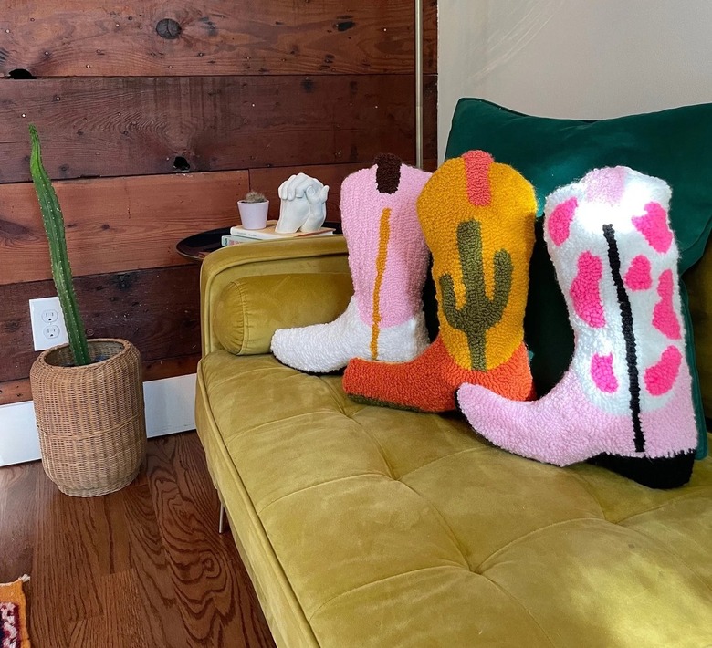 Three tufted cowboy boot pillows on a sage green couch. One is a pink and white bow pattern
