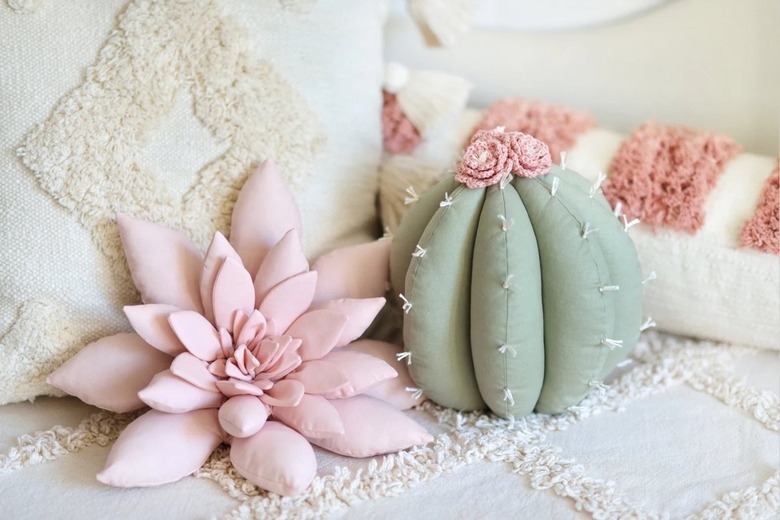A pink succulent-shaped pillow and green cactus-shaped pillow with pink flowers on top. They are on a white bed with lines that have a fuzzy texture.
