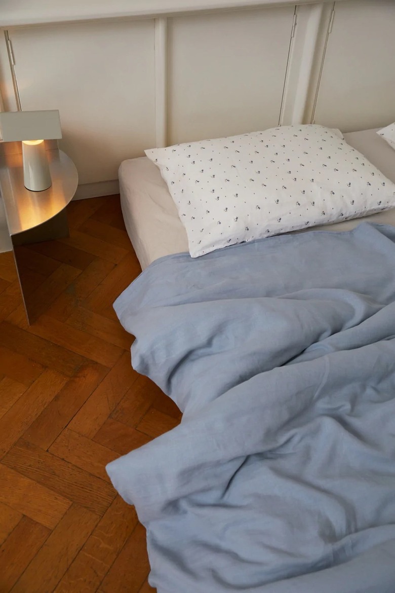 White floral pillow and a light blue duvet on a mattress on the hardwood floor.