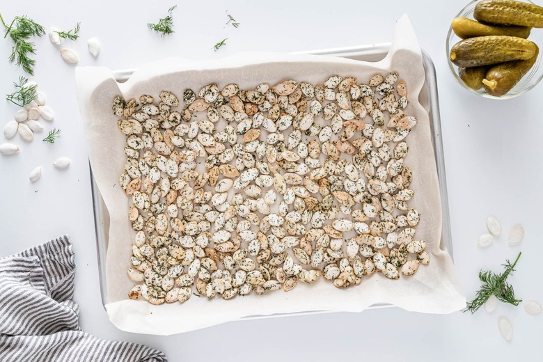 A parchment-lined silver baking sheet covered in the dill pumpkin seeds on a white counter. There is also a small bowl of dill pickles