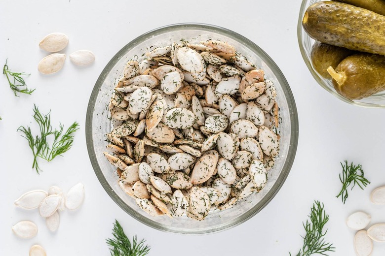 A small glass bowl of pumpkin seeds and the olive oil mixture now coated into dried herbs on a white countertop.