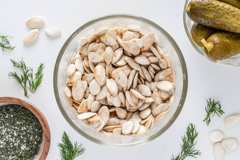 A small glass bowl of pumpkin seeds coated in the olive oil mixture on a white counter.