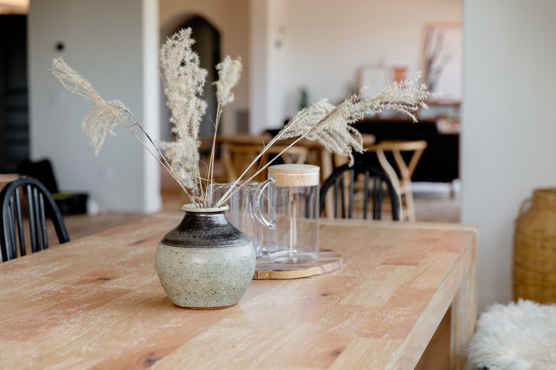 Light wood tabletop with a grey and black ceramic vase with stalks of wispy grass in it. Behind the vase