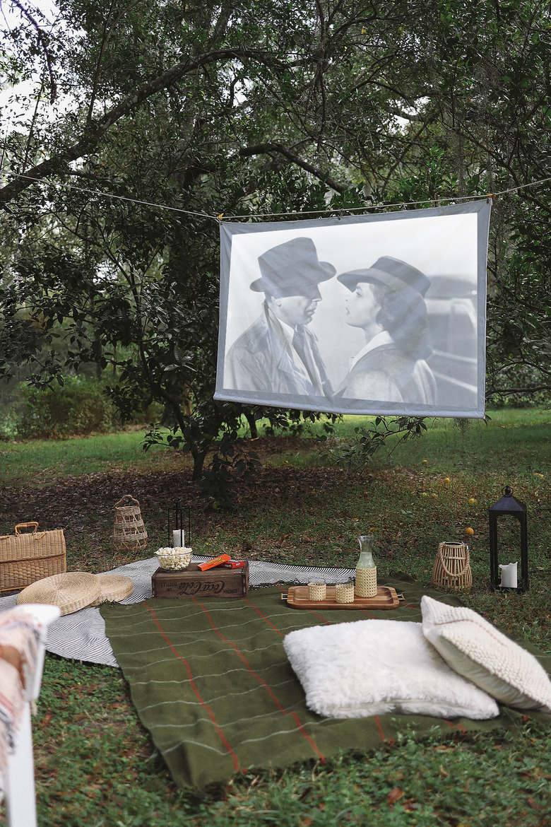 DIY portable projector screen hanging outdoors between trees with blankets and snacks set up below