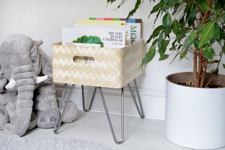 Light tan book bin with metal legs and picture books in a white room next to elephant stuffed animal and medium-sized plant