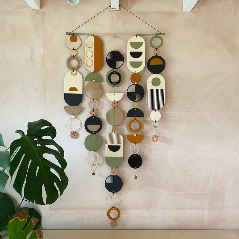Wall hanging with geometric shapes in neutral tones. There is a plant in the corner.