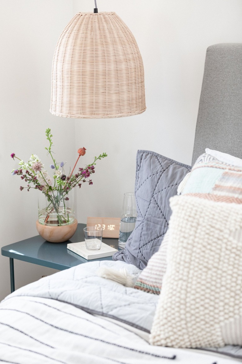A wicker pendant lamp over a blue nightstand with flowers and bed with pillows