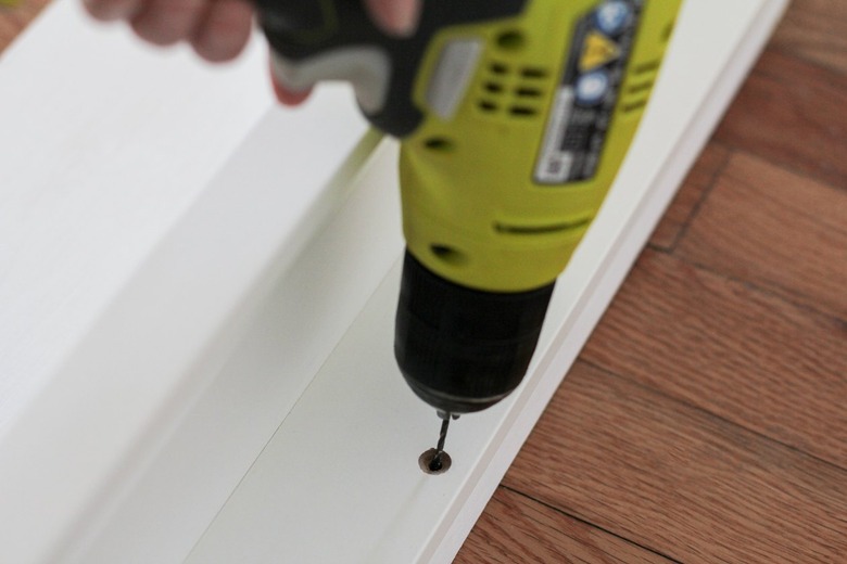 Hand screwing gold screw into white board atop wooden flooring