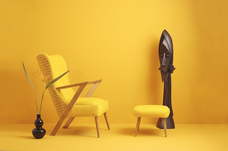 A bright yellow armchair and ottoman on a yellow background next to a large vertical wood African mask.