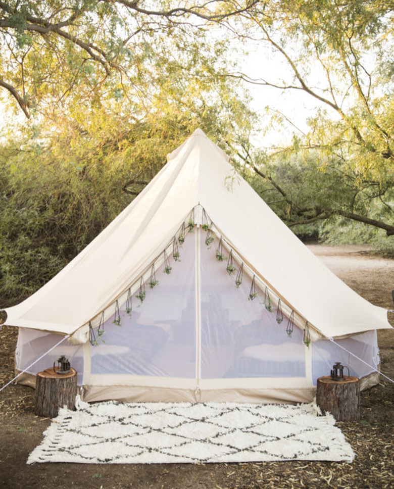 A photo of a white tent with mesh sides and tassels in the woods