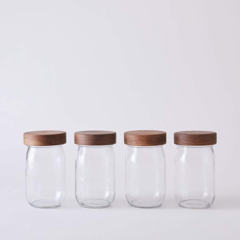 Four clear glass spice jars with wood lids in a row over a white background.