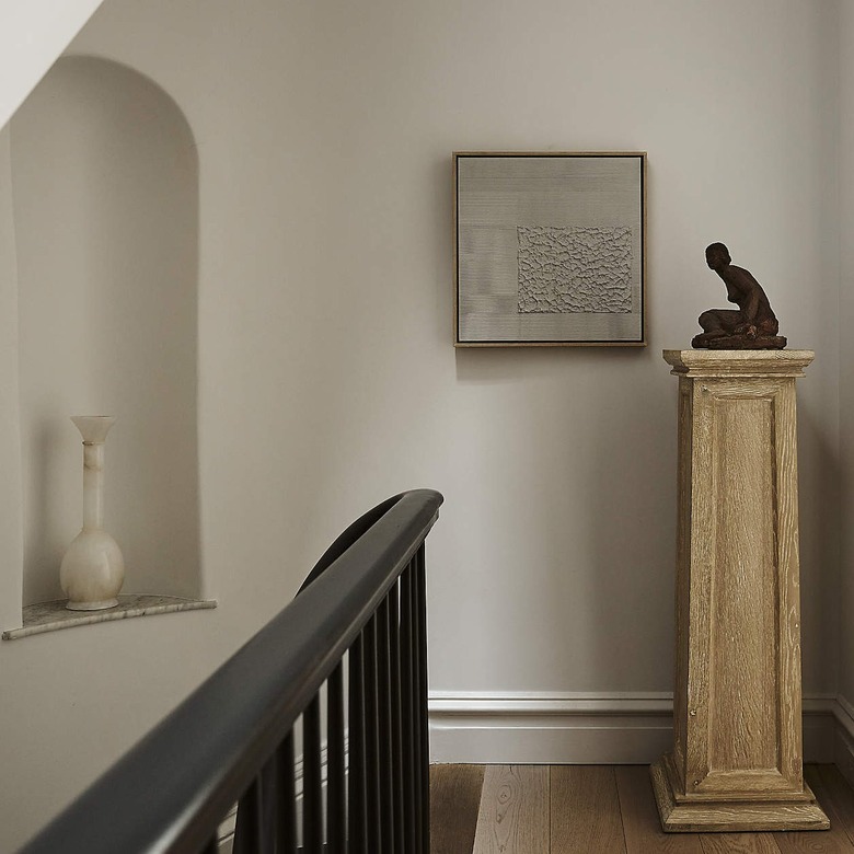 A piece of gray textured art on a white wall next to a beige column pedestal.