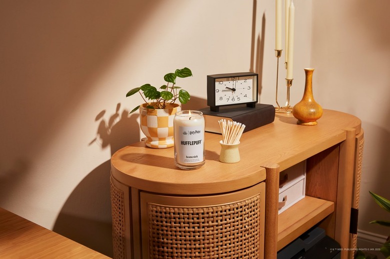 Homesick's Hufflepuff candle on a wood and rattan side table with a clock and candles in gold holders.