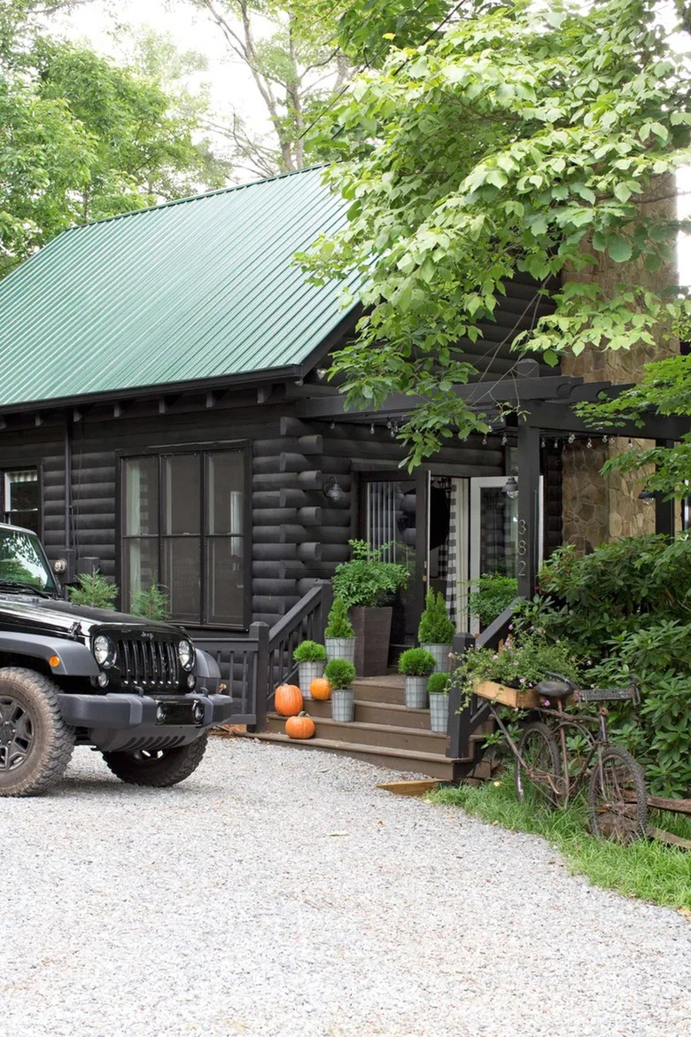 A black log cabin-style house has a green metal roof. There are pumpkins on the stairs and a Jeep parked in front of the pea gravel driveway.