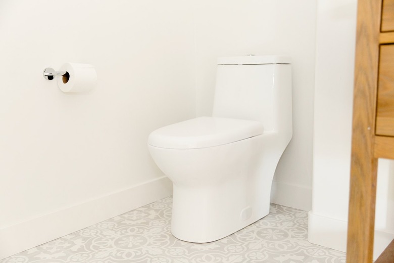 A white toilet in a bathroom with white walls and a white and grey tiled floor. A roll of toilet paper is on the wall next to the toilet