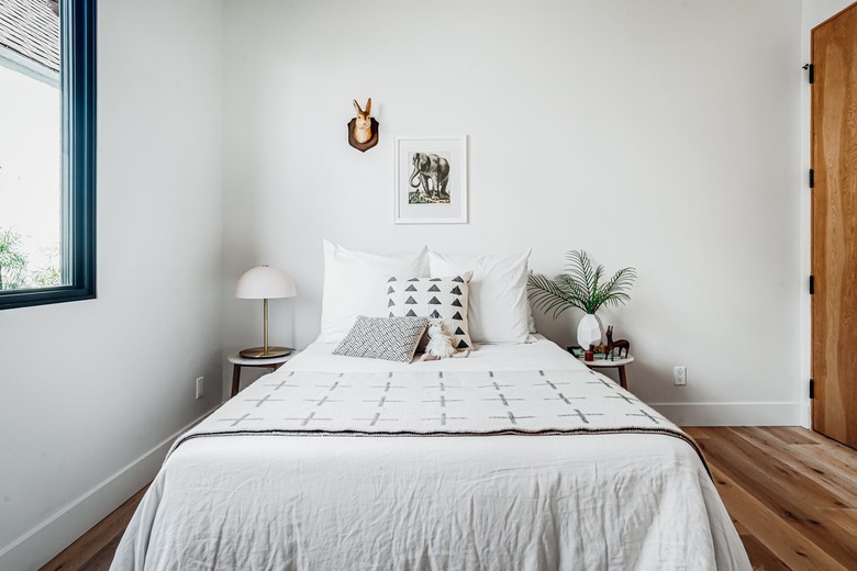 White bedding with patterned pillows and a stuffed animal. A white half dome table lamp. A geometric vase with palm leaves