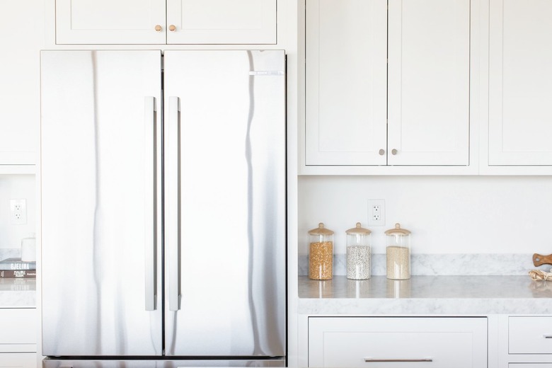 Double doors of a stainless steel refrigerator with white cabinets overhead. Next to it