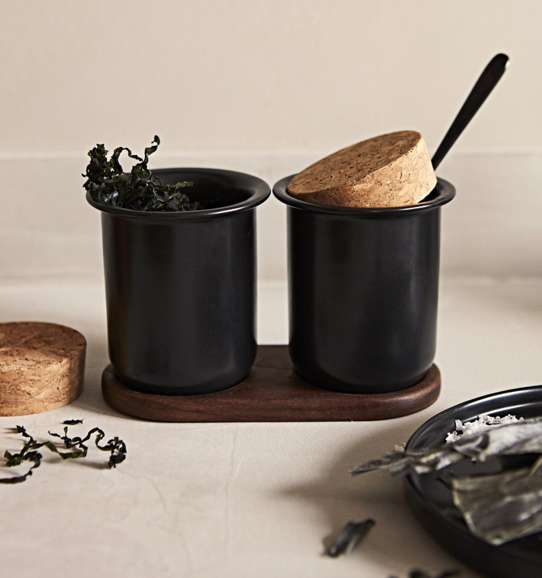 Two black cups on a wooden platform on a countertop with cork stoppers and silverware sticking out.