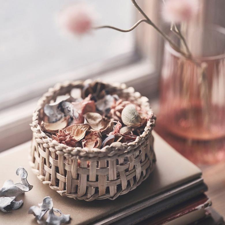 Woven wooden basket filled with potpourri next to a vase with pink flowers in front of a window
