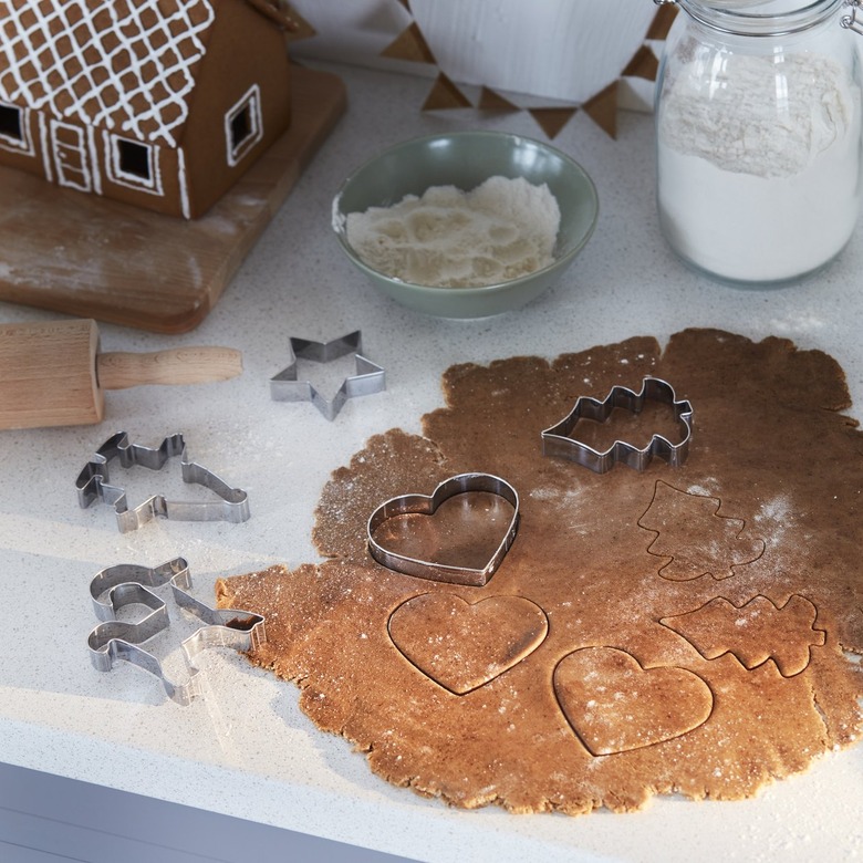 Christmas tree and heart-shaped cookie cutters placed on cookie dough that is spread on a white countertop. There is a gingerbread house and sugar in the background.
