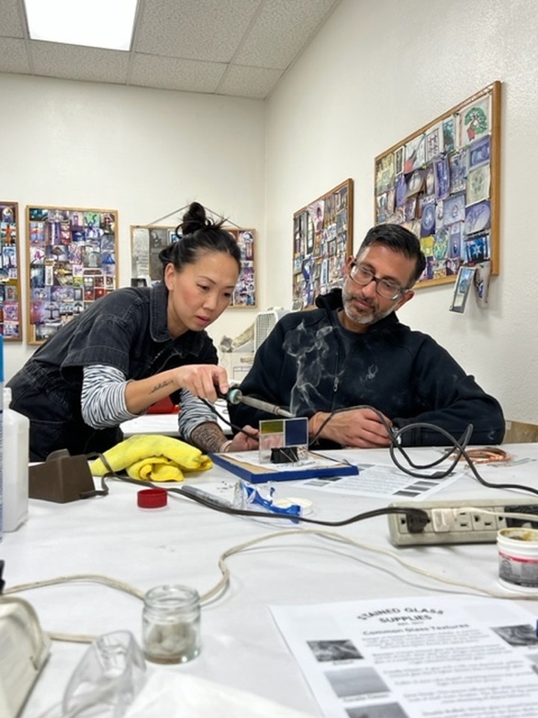 Asian woman with black hair pulled back is wearing a black top with blue and white striped sleeves underneath. She is demonstrating how to cut glass to a man with medium brown skin