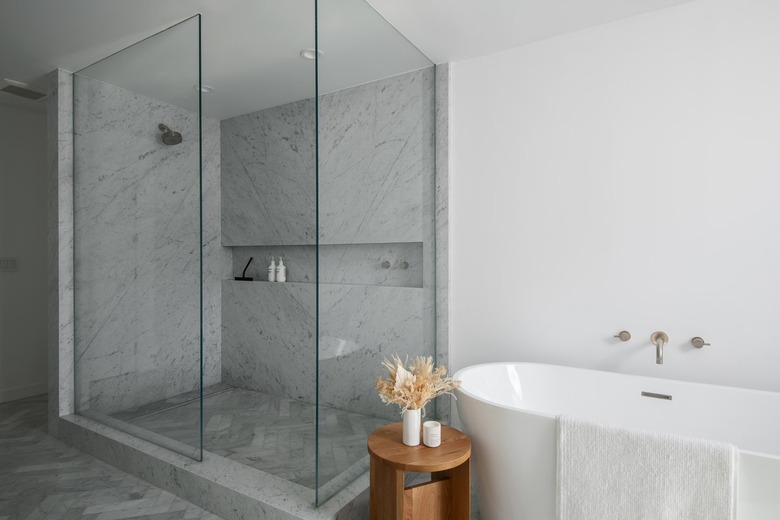 A white bathroom with a white and gray marble standing shower surrounded by glass
