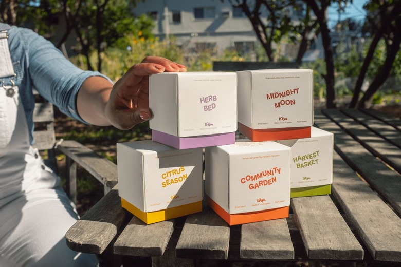 Jessica White placing her hand on a pile of five boxes of Loam Candles on a wood slat table in an outdoor garden.