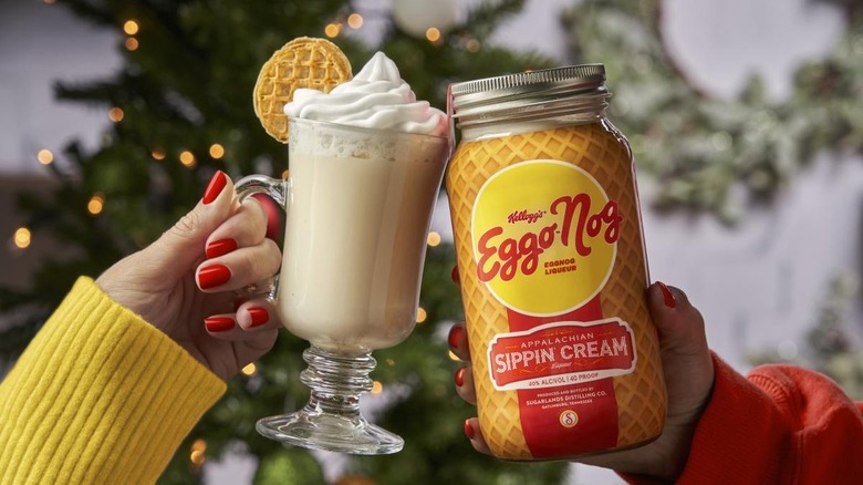 A glass and a jar of Eggo Nog toast in front of a lit Christmas tree: The glass on the left is a tall