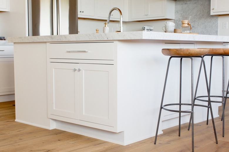 A row of bar stools with wooden seats and black metal legs in front of a white kitchen island with a marble countertop. Visible on the countertop is a chrome faucet