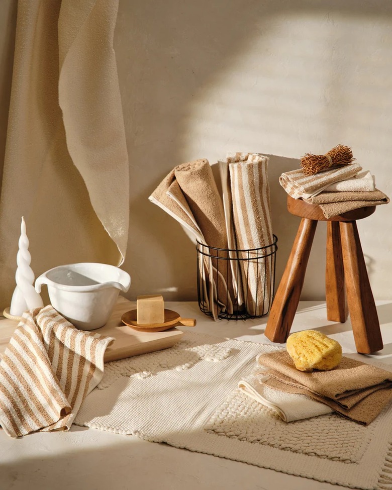 Beige and white striped and solid towels are folded and rolled in a bathroom setting with wood stool and tray