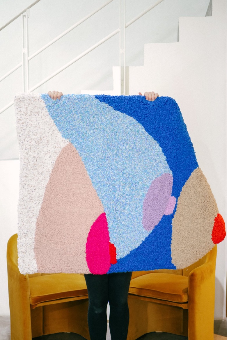 Colorful rug of three breasts and nipples being held by two hands with white staircase and rust chairs in the background