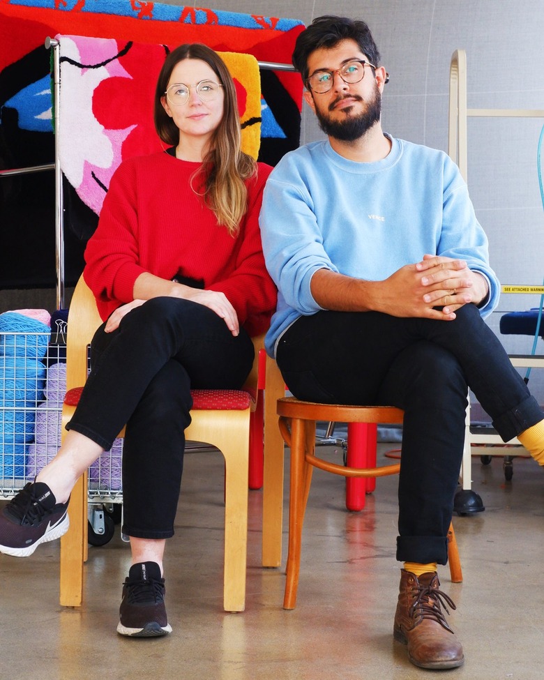 A woman with long brown hair and long-sleeved red sweater and a bearded man with short brown hair and light blue sweater both wear glasses and black jeans sit next to each other on different chairs