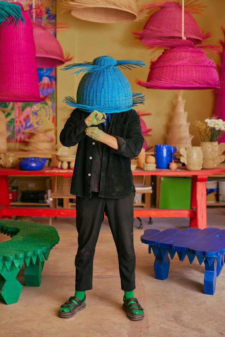 A man dressed in black holding a bright blue wicker basket over his head in front of a studio composed of a work station