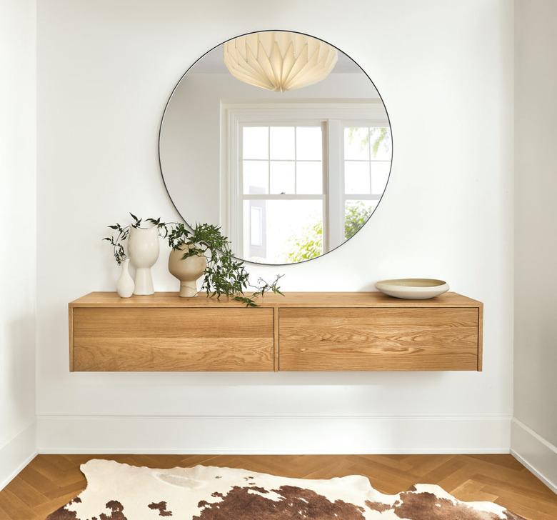 Minimalist entryway design with circular mirror over floating wood cabinet on white wall with cowhide below.